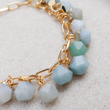 A close-up of a gold chain bracelet featuring faceted beads in pale icy blue, white, and mint green, resting on a textured light beige surface.